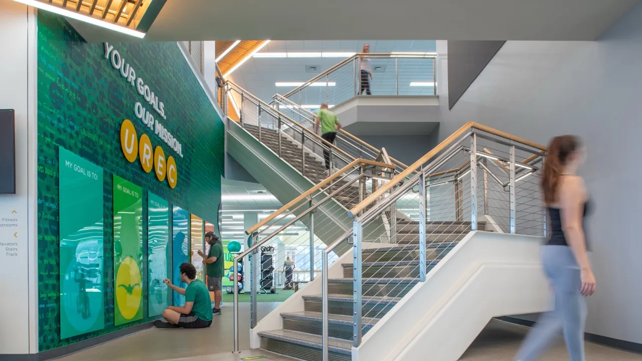 Interior of the University Recreation Center, there is an interactive goal wall to the left and a staircase in the center. people walk by